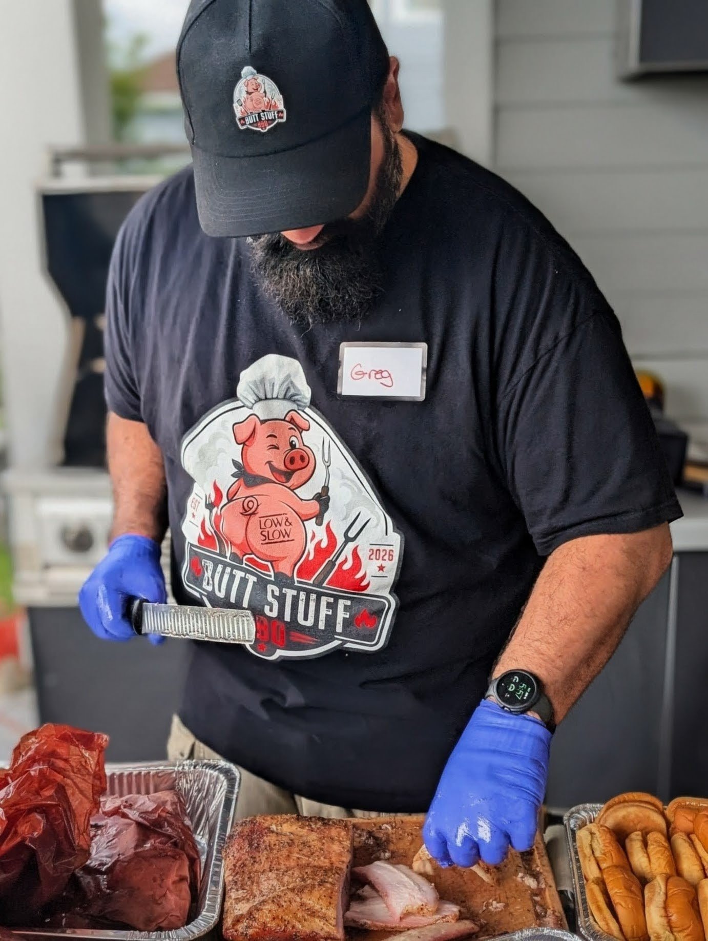 Greg, the founder of Butt Stuff BBQ, in a branded cap and shirt slicing smoked pork over trays of meat and buns at his backyard cookout.